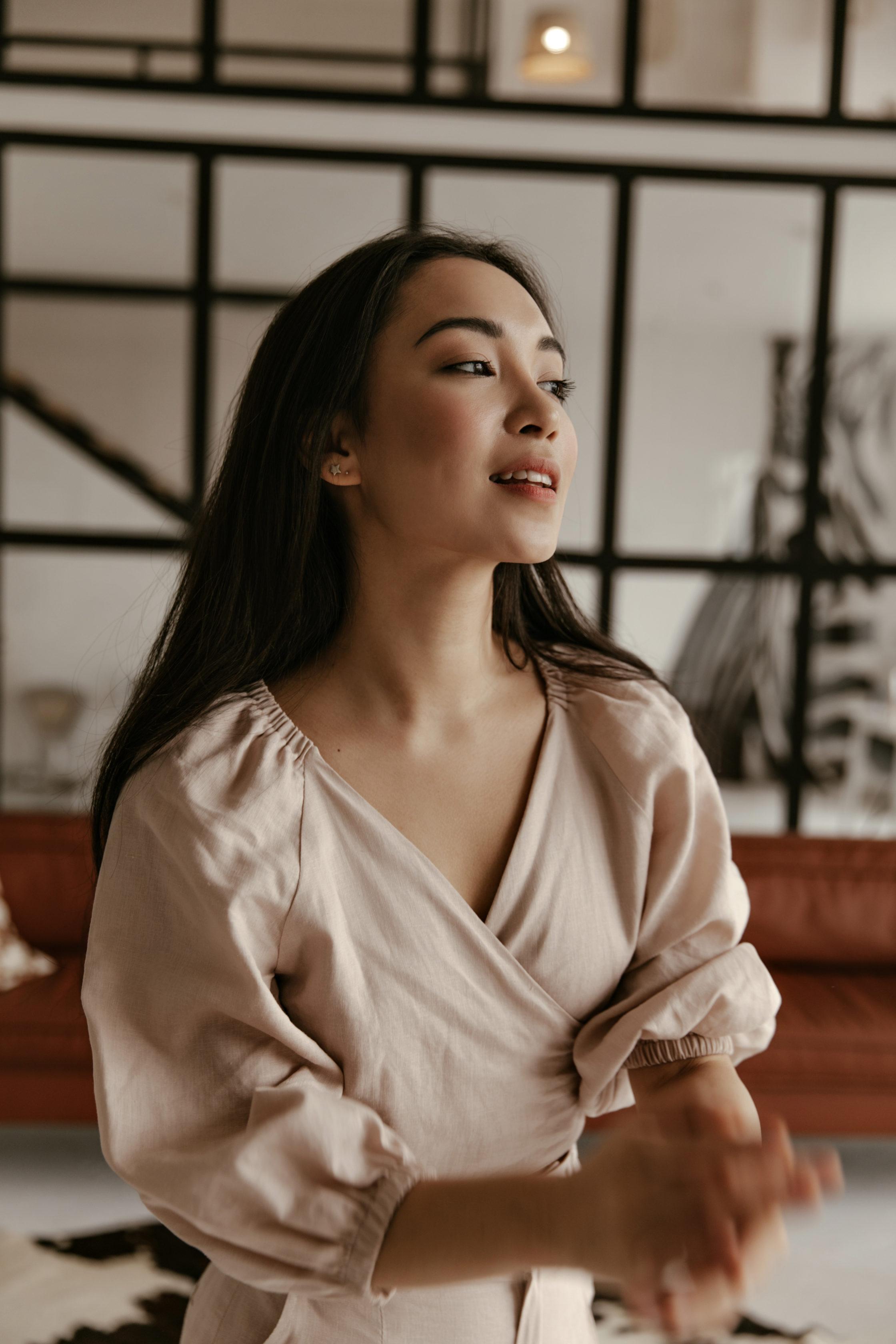 Calm self-confident young woman in beige linen blouse smiles and looks away. Portrait of brunette Asian woman in cozy living room.
