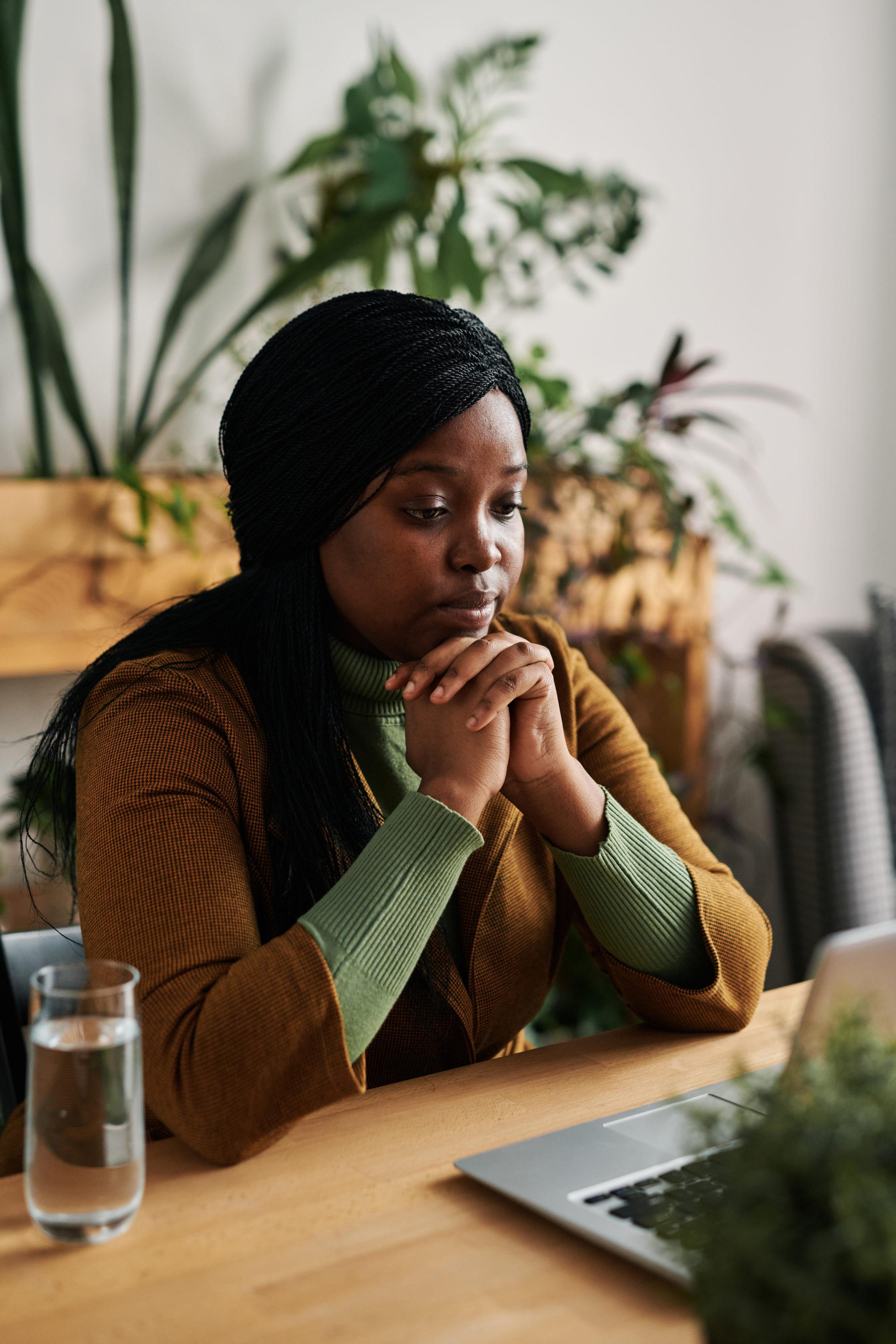 Young serious psychoanalyst listening to online patient while sitting by workplace in front of laptop and keeping her hands by chin